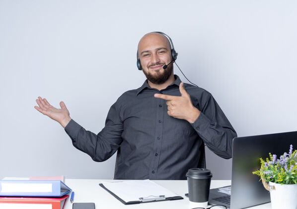 年轻高兴的年轻秃头呼叫中心男子戴着耳机坐在办公桌旁的工作工具显示空手 并指着它孤立的白色背景中心工具手