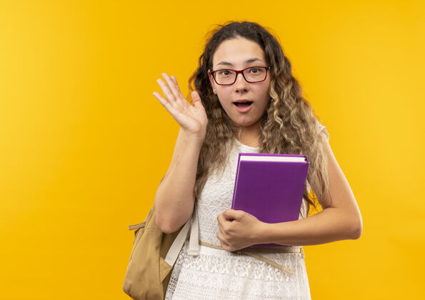 黄色令人印象深刻的年轻漂亮的女学生戴着眼镜 背着包拿着一本书 显示空的手孤立在黄色抱印象包