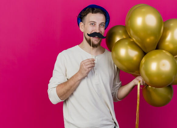 假有趣的微笑年轻英俊的斯拉夫党的家伙戴着党的帽子 拿着气球和假胡子坚持以上的嘴唇看前面的粉红色墙壁上的复制空间孤立人胡子嘴唇