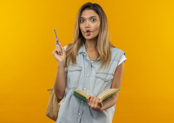抱印象深刻的年轻漂亮的学生女孩穿着背袋举行开放的书和笔孤立的橙色笔包年轻