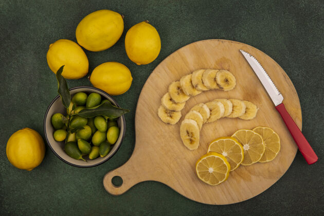 餐新鲜水果片的俯视图 如香蕉和柠檬 放在木制厨房板上 刀子上放着金盏花 碗里的柠檬被隔离在绿色的背景上水果板饮食