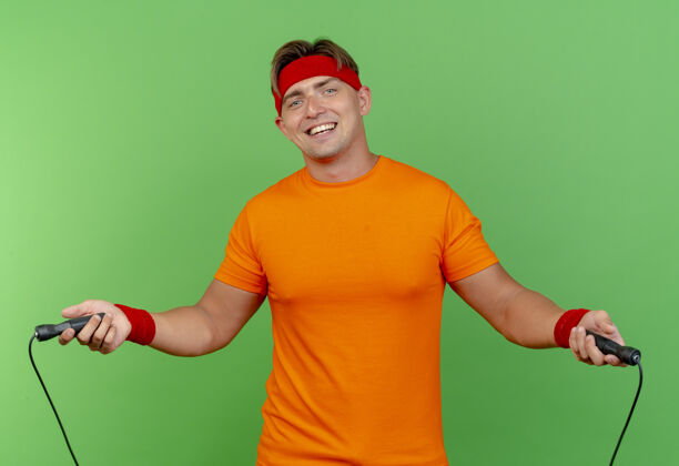 英俊快乐的年轻英俊的运动男子戴着头带和腕带跳绳隔离在绿色穿头带绿色