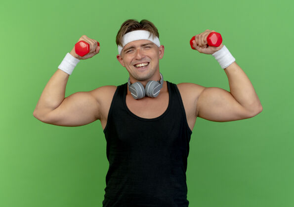 欢乐快乐的年轻帅气的运动型男人戴着头带和腕带 脖子上戴着耳机 举着绿色的哑铃耳机运动腕带