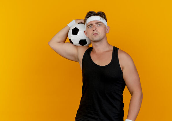 相机年轻帅气的运动型男人戴着头带和腕带 肩上扛着足球 看着隔离在橙色背景上的相机 还有复制空间帅气头带运动