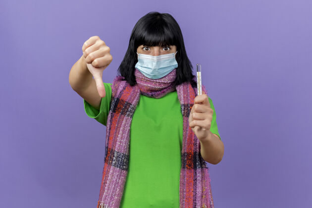 年轻人印象深刻的年轻病妇女戴着口罩和围巾拿着温度计看着前面显示拇指向下孤立的紫色墙壁与复制空间姿势面具放下