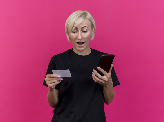 金发印象深刻的中年金发斯拉夫妇女手持手机和信用卡看着卡隔离在粉红色墙上的复制空间衣服电话公民