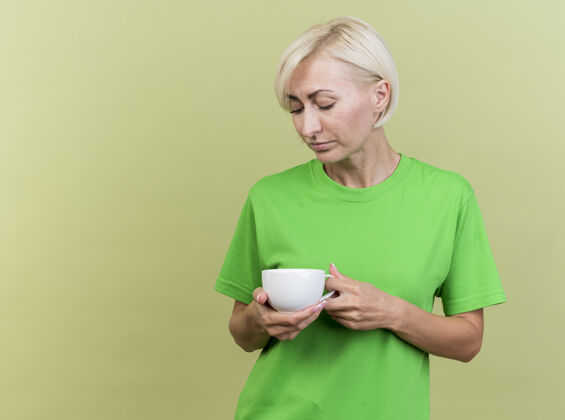 站着悲伤的中年金发斯拉夫女人拿着一杯茶 看着隔离在橄榄绿墙上的复制空间杯子橄榄女人