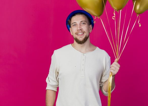 穿着面带微笑的年轻英俊的斯拉夫党人戴着党的帽子拿着气球看着前面孤立的粉红色墙上的复制空间抱着人姿势