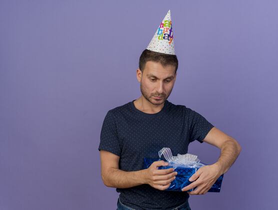 失望失望的帅哥戴着生日帽拿着礼盒孤立地放在紫色的墙上 有复制空间生日举行感情