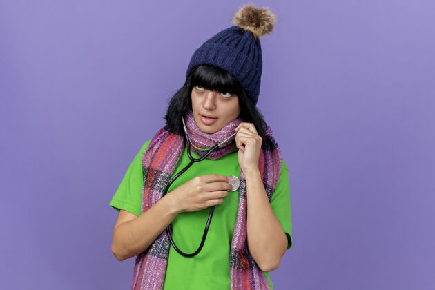 女孩专注的年轻病女人戴着冬天的帽子 戴着围巾 用听诊器看着一边听着她的心跳 隔离在紫色的墙上 留着复制空间听诊器感觉人
