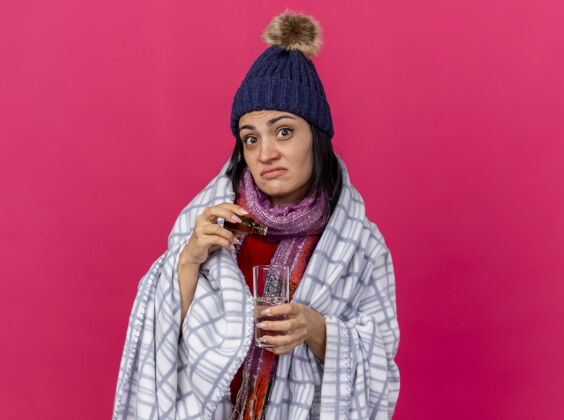 冬天印象深刻的年轻生病的妇女戴着冬天的帽子和围巾在格子布包裹加入药剂到水玻璃看前面的粉红色墙壁与复制空间隔离水感情医疗