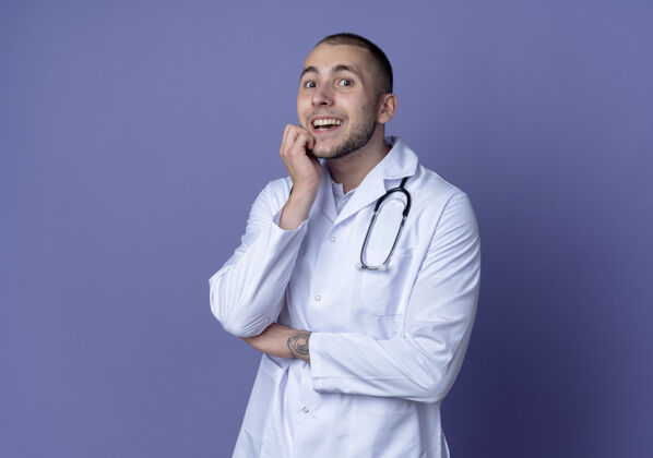 印象印象深刻的年轻男医生穿着医用长袍和听诊器把手放在下巴和肘部隔离在紫色肘部长袍手