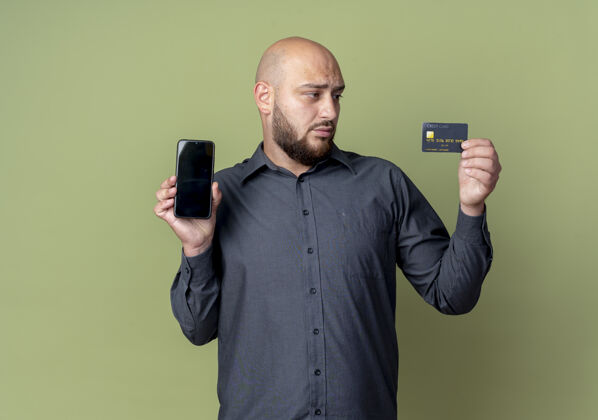电话年轻的秃头呼叫中心男子手持手机和信用卡 看着卡孤立的橄榄绿背景与复制空间秃头年轻手机