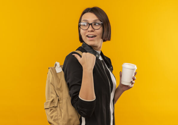 塑料快乐的年轻学生女孩戴着眼镜 背着书包站在侧视图中 拿着塑料咖啡杯 指着身后孤立的橙色眼镜轮廓站着