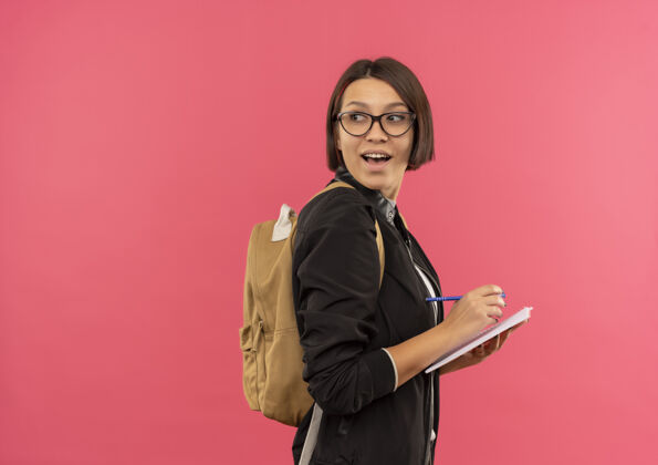 粉红色快乐的年轻学生女孩戴着眼镜 背着书包站在侧视图中 拿着钢笔和记事本 看着身后孤立的粉红色包眼镜垫
