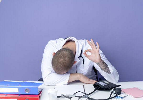 医生疲惫的年轻男性医生穿着医用长袍和听诊器坐在办公桌旁 把头放在办公桌上 在紫色背景上做着“ok”标志工作听诊器长袍