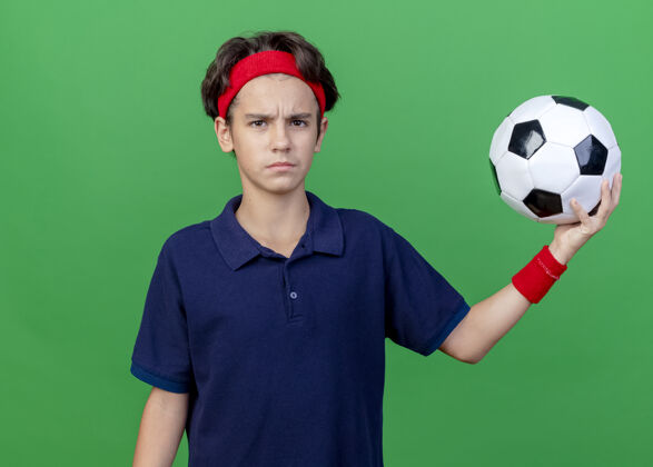年轻皱眉的年轻英俊的运动男孩戴着头带和戴着牙套的护腕拿着足球看着绿色墙壁上孤立的前方男孩头带戴