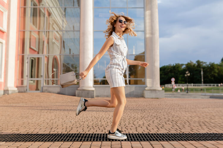 女性年轻迷人的时尚女子穿着运动鞋在城市街道上奔跑跳跃有趣夏天时尚风格的白色连衣裙戴着太阳镜和手提包城市城市优雅