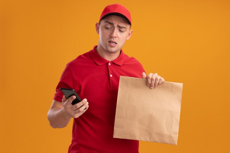 食物年轻的送货员穿着制服 戴着帽子 手里拿着一个纸制食品包 看着手机 把手机孤立在橙色的墙上帽子送货年轻