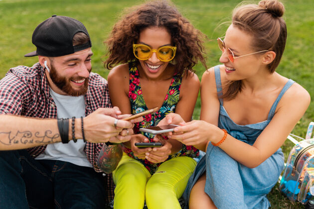 无线快乐的年轻朋友们坐在公园里用智能手机微笑 男人和女人一起玩音乐微笑耳机