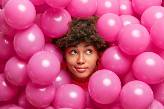 满意美籍黑人妇女喜欢在粉红色的气球中摆姿势 沉思地看着旁边的人 为聚会做计划庆典女性发型