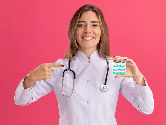 年轻面带微笑的年轻女医生穿着医用长袍 手持听诊器 指着隔离在粉红色墙上的药片女性医生长袍