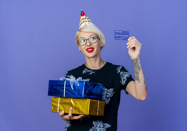 礼物微笑的年轻金发派对女孩戴着眼镜 戴着生日帽 拿着礼品盒和信用卡 看着紫色背景上有复制空间的隔离卡微笑女孩信用卡