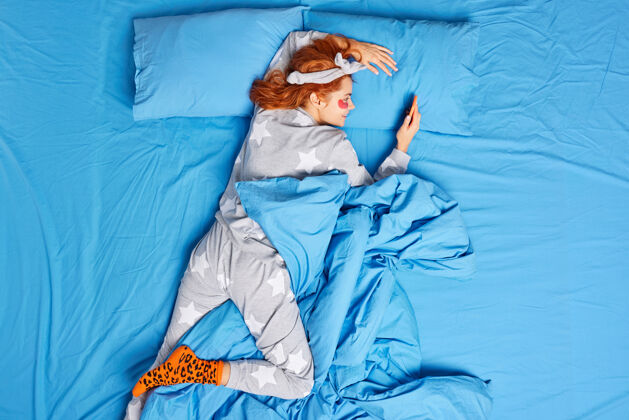 姜懒洋洋的姜女穿着舒适的睡衣 睡后在眼睛下敷上水凝胶贴片 以减少皱纹和浮肿躺在床上蓝色床单上用手机聊天室内睡衣休闲