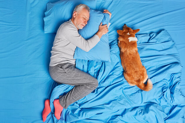 休闲老年人的男人狗主人安详地睡在一起 在床上摆宠物姿势 穿着睡衣和袜子躺在柔软的枕头上 看到甜蜜的梦成熟的胡须男人在卧室休息人们睡觉的概念夜晚男性人