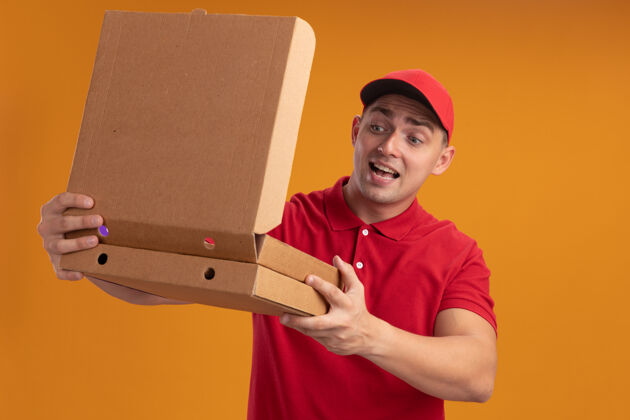 制服快乐的年轻送货员穿着制服 戴着帽子 看着隔离在橙色墙上的披萨盒年轻穿欢乐