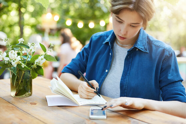 笔记用智能手机做笔记的女人一个年轻女人写在笔记本上的户外肖像莫比尔笔记本钢笔