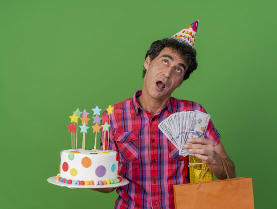 蛋糕印象深刻的中年白种人聚会男子戴着生日帽拿着生日蛋糕纸袋礼包和钱抬头看绿色背景上孤立纸印象聚会