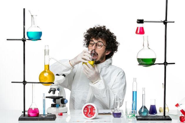 化学正面图身着特殊套装的年轻男科学家坐在白墙上 喝着饮料饮用水烧杯成人
