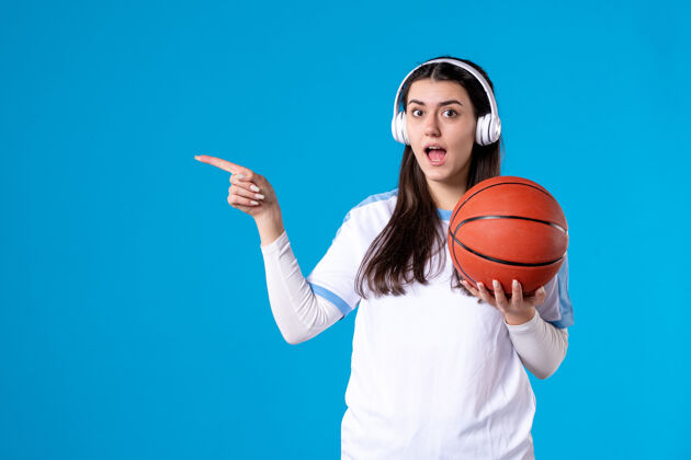 肖像正面图：戴着耳机拿着篮球的年轻女性站在蓝色的墙上正面人物耳机