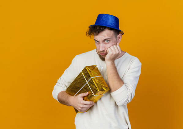橙色可疑的年轻英俊的斯拉夫党人戴着党的帽子拿着礼品盒把手放在脸上看着相机孤立在橙色背景与复制空间脸帽子怀疑