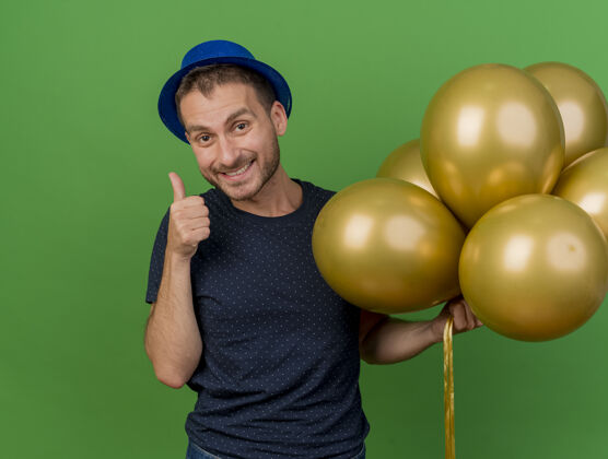复制面带微笑的白人帅哥戴着蓝色派对帽竖起大拇指 拿着氦气球隔离在绿色背景上 留有复制空间男人向上空间