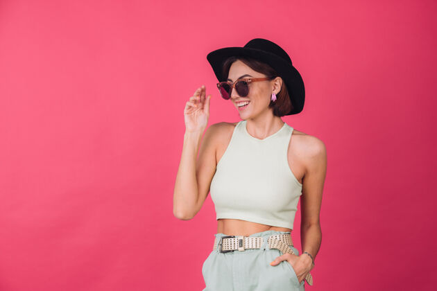 夏天戴着帽子和太阳镜的漂亮时髦女人在粉红色的墙上摆姿势华丽帽子时尚