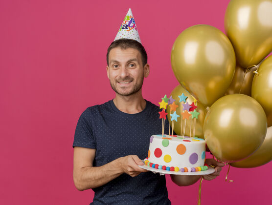 微笑微笑英俊的白人男子戴着生日帽拿着生日蛋糕和氦气球隔离在粉红色背景与复制空间气球帽子空间
