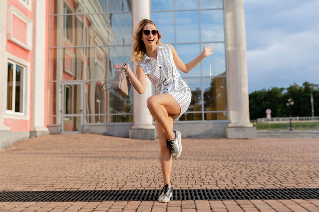 华丽年轻迷人的时尚女子穿着运动鞋在城市街道上奔跑跳跃有趣夏天时尚风格的白色连衣裙戴着太阳镜和手提包城市服装女人