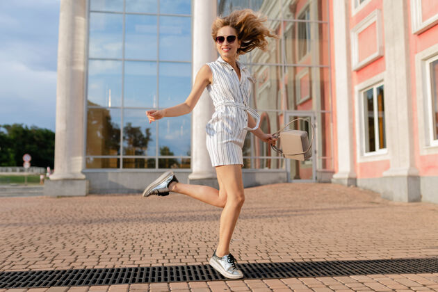 魅力年轻迷人的时尚女子穿着运动鞋在城市街道上奔跑跳跃有趣夏天时尚风格的白色连衣裙戴着太阳镜和手提包街道城市女孩