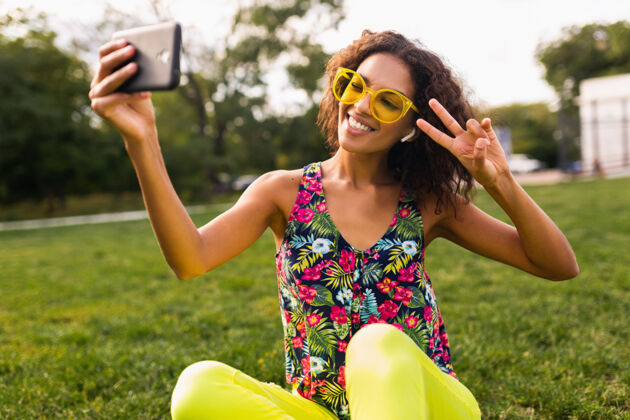女人年轻时尚积极的黑人女性 自拍 戴着无线耳机听音乐 在公园里玩得很开心 夏日时尚 五颜六色的时髦装扮时尚眼镜兴奋