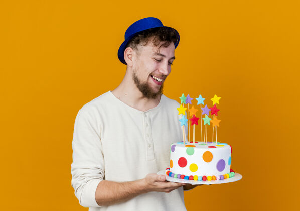 年轻人快乐的年轻帅哥斯拉夫党家伙戴着党的帽子举行 看着生日蛋糕与星星孤立在橙色背景与复制空间橙色斯拉夫人太空