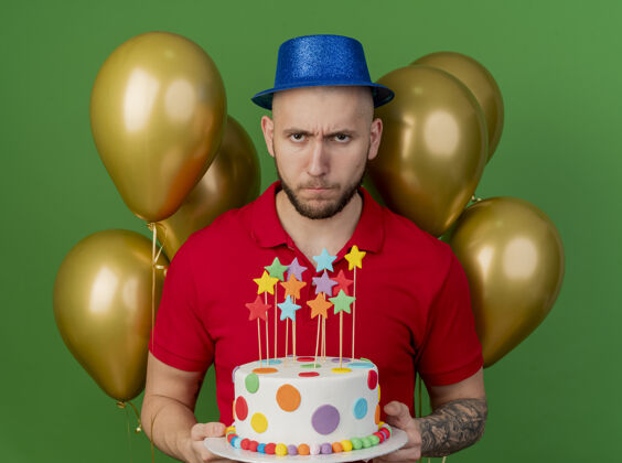 皱眉皱眉的年轻帅哥斯拉夫党人戴着党帽站在气球前拿着生日蛋糕看着相机隔离在绿色背景气球站着帽子