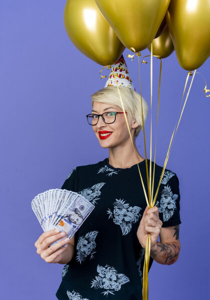 派对微笑的金发派对女郎戴着眼镜 戴着生日帽 手里拿着气球和钱 看着紫色背景上孤立的相机气球紫色生日