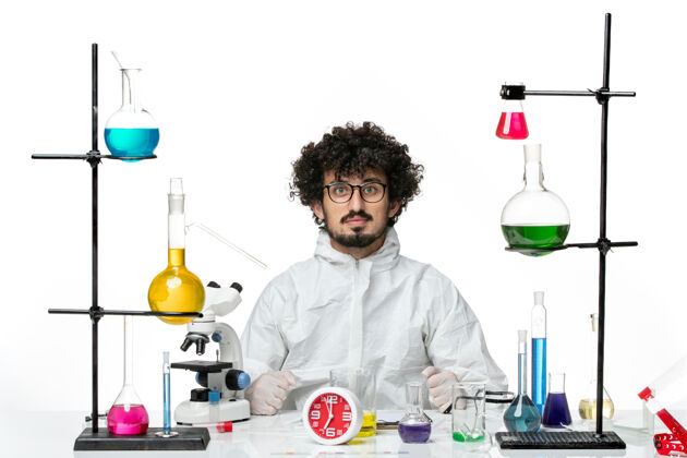 烧杯正面图身着特殊套装的年轻男科学家坐在白墙上 用不同的解决方案化学化学玻璃