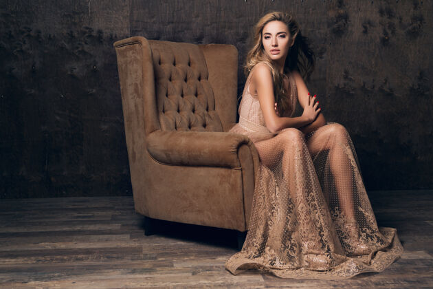 皮肤穿着闪亮蕾丝晚礼服的美丽性感模特坐在米色的椅子上女人人室内