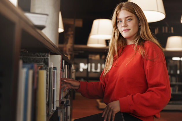 考试漂亮的红发女孩 大学生从图书馆或书店的书架上拿起一本书 对着镜头微笑成人书架青少年