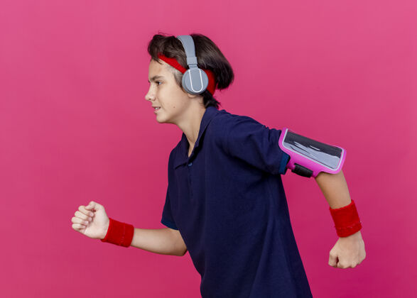 年轻年轻帅气的运动男孩戴着头带 腕带和耳机戴着牙套的电话臂带笔直地奔跑着保持双手在空气中与深红色背景隔离腕带跑步电话