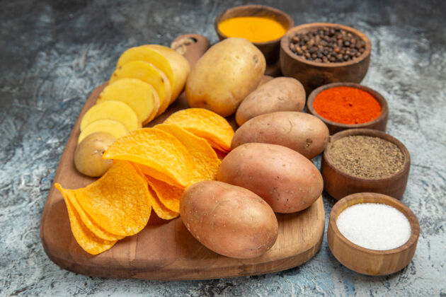 水果脆薯片和生土豆放在木制砧板上 不同的香料放在灰色的桌子上香料营养食品
