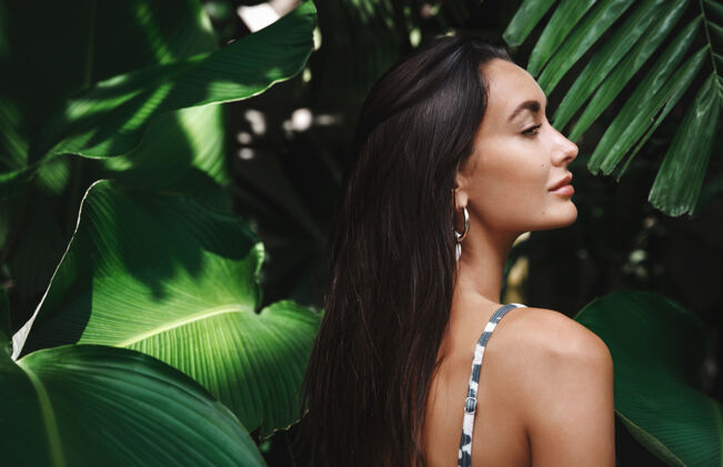 热带美丽的深褐色女人的后视图 穿着比基尼 站在绿叶的侧面树叶丛林比基尼